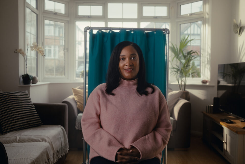 A woman in a pink jumper sat in her living room. There is a portable hospital curtain staged behind her.